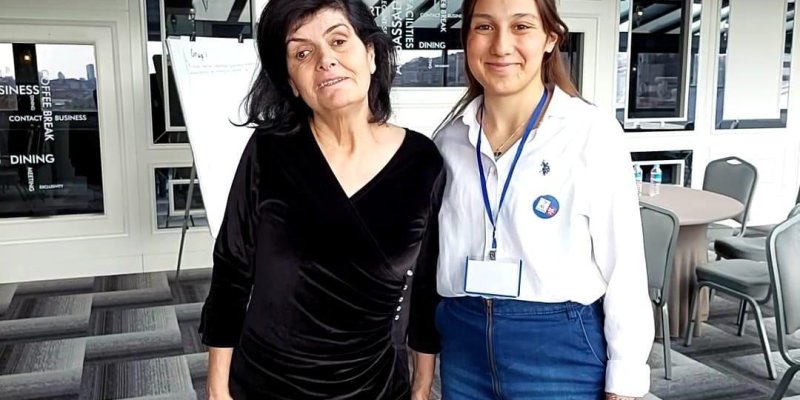 İki kadın birlikte duruyor. Sol taraftaki kadın siyah bir elbise giymiş ve saçları omuzlarına kadar uzun. Sağ taraftaki kadın ise mavi bir etek ve beyaz bir gömlek giymiş, kimlik kartı asılı ve saçları uzun ve açık renk. Arka planda gri tonlarda bir halı, beyaz duvarlar ve kapalı bir mekanın içi görülüyor.