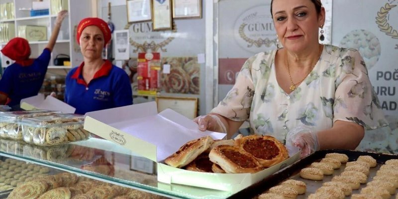 Tezgâhın arkasında bir pastane veya fırın içinde çalışan üç kadın ve tezgâhın üzerinde çeşitli hamur işleri. Önde, açık renk çiçekli bluz giymiş, sarı saçları arkadan toplanmış orta yaşlı bir kadın, beyaz karton kutuyu tezgâhın üzerine doğru uzatıyor; kutunun içinde üstü kıymalı ve biberli görünen, yuvarlak, kenarları kabarık küçük pideler sıralı duruyor. Kadının elinde şeffaf hijyen eldiveni ve boynunda ince bir zincir kolye; yüzünde hafif ciddi ama sıcak bir ifade. Sağ tarafta, tezgâhın üzerin