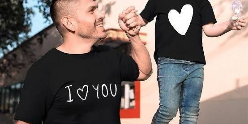 ir adam ve küçük bir kız çocuğu el ele tutuşmuşlar. Adamın üzerinde "I ♥ YOU" yazılı siyah bir tişört, küçük kızın üzerinde büyük beyaz bir kalp bulunan siyah bir tişört var. Kız çocuğu kot pantolon ve beyaz spor ayakkabı giymiş. İkisi de mutlu görünüyor ve adam kıza sevgi dolu bir şekilde bakıyor. Arka planda ağaçlar ve bir bina var. Kız çocuğu elinde bir dondurma tutuyor.