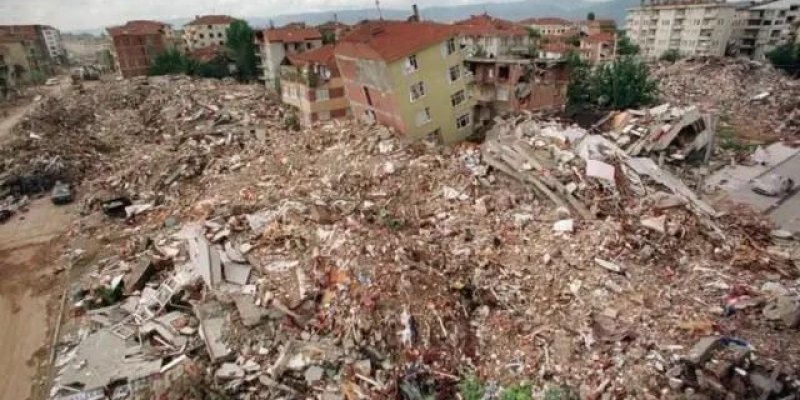 Deprem sonrası yıkıntılarla kaplı geniş bir şehir manzarası. Kare boyunca neredeyse tamamen tuğla, beton, demir ve moloz yığınları görülüyor; çok sayıda bina tamamen çökmüş. Orta kısımda birkaç katı ayakta kalmış, sarı ve pembe tonlarında, kırmızı çatılı bir apartman kısmen eğilmiş şekilde; yan cepheleri açılmış, iç odalar dışarıdan görünür hâle gelmiş. Sağ tarafta büyük bir bina bloğunun katları üst üste çökmüş, beton plakalar ve demir kolonlar yan yatmış olarak molozun üzerinde uzanıyor. Sol a