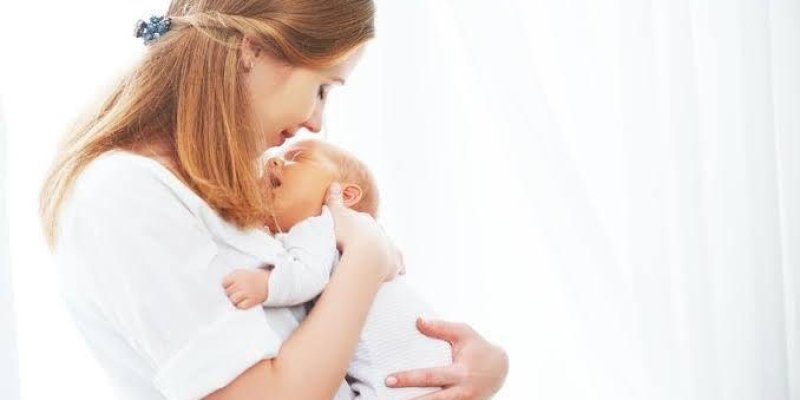 Bir anne ve kucağında tuttuğu bebek. Anne, bebeğini sevgiyle göğsüne yaslamış ve ona gülümsüyor. İkisi de beyaz giysiler giymiş. Arka plan çok aydınlık ve beyaz. Anne ve bebek arasında yakın ve sevgi dolu bir bağ hissediliyor.