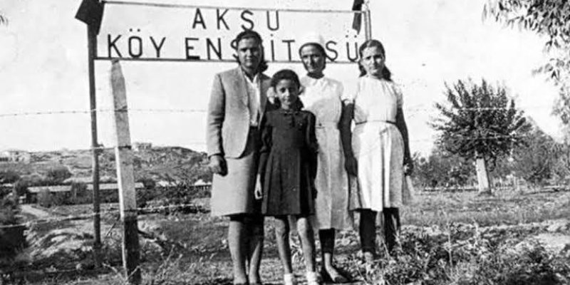 Bu siyah-beyaz fotoğrafta dört kadın bir arada poz veriyor. Arkalarında "AKSU KÖY ENSTİTÜSÜ" yazılı bir tabela bulunuyor. Kadınlar, kırsal bir alanda, açık havada duruyorlar. Arka planda ağaçlar ve ufukta tepeler görülüyor. Biri ceket ve etek, diğerleri elbise giymiş.