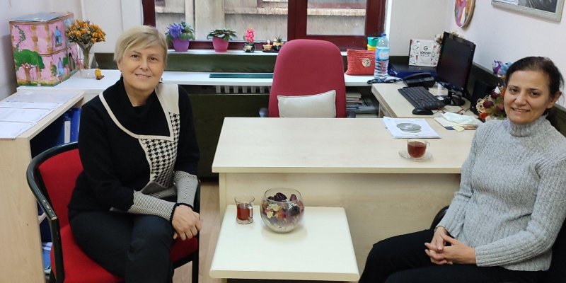 Pencere önündeki ve vazodaki çiçeklerin olduğu ofiste çalışma masasının ön tarafında iki kadın gülümsüyor. Soldaki kırmızı sandalyede kısa sarı saçlı, siyah kıyafetli, bacak bacak üstüne atmış, elleri de birbirinin üzerinde oturuyor. Sağdaki, ofis sandalyesinde gri kazaklı, siyah pantolonlu koyu renk saçlarını toplamış ellerini birbirine kavuşturmuş. Önlerinde çayları var.