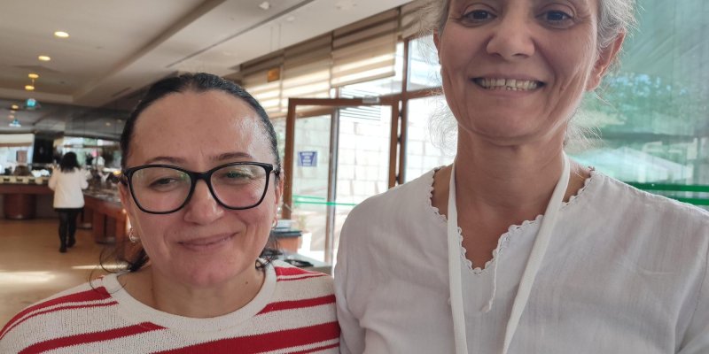 İki kadın omuz omuza duruyor ve kameraya doğru gülümsüyorlar. Sol taraftaki kadın koyu saçlı, gözlüklü ve beyaz zemin üzerine kırmızı çizgili bir kazak giymiş. Sağ taraftaki kadın ise açık gri saçlı, beyaz bir bluz giymiş ve makyajsız doğal bir görünüme sahip. Arka planda açık renkli bir restoranın iç mekanı ve camdan dışarıya bakan bir pencere görülüyor.