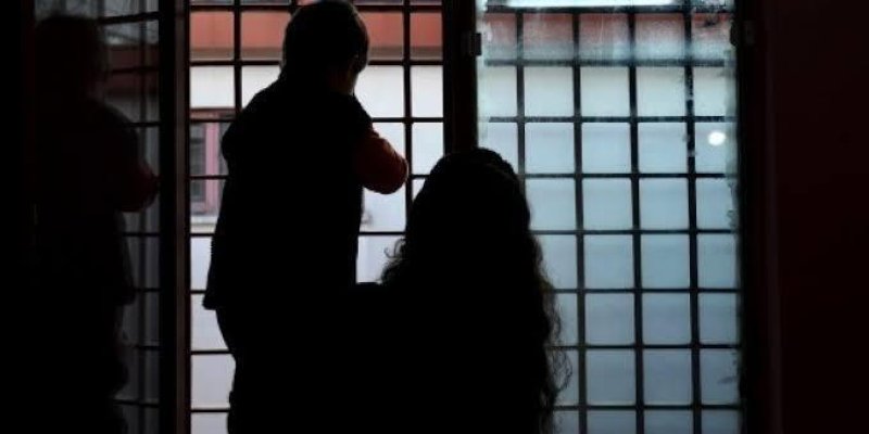 Pencere parmaklıklarının önünde dışarıya dönük duran iki çocuk ve bir yetişkinin silüeti. Ortada, kolları hafifçe yukarıda, yüzü dışarıya dönük bir çocuk silüeti; omuz hizasında kısa saçlı, üstünde koyu renk yelek ya da mont, kollarında daha açık tonda uzun kollu bir kıyafet. Çocuğun önünde, yere daha yakın konumda, uzun ve dalgalı saçları bel hizasına kadar inen, çömelen başı dışarıya dönük bir yetişkin silüeti. Solda, pencerenin kenarına yakın, boyu daha kısa başka bir çocuk silüeti duvara ya
