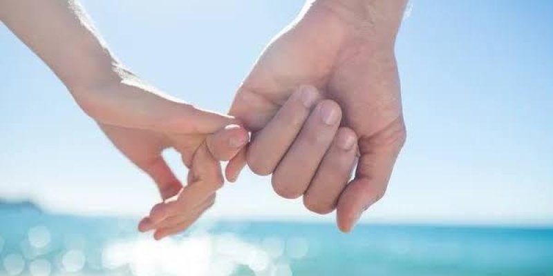 Birbirini tutan iki el. Bir kadın ve bir erkeğin elleri birbirine kenetlenmiş. Arka planda deniz ve gökyüzü var; hava güneşli ve parlak. Ellerin odakta olduğu bu karede, denizin maviliği ve güneş ışığının yansımaları flu şekilde arka planda kalıyor.