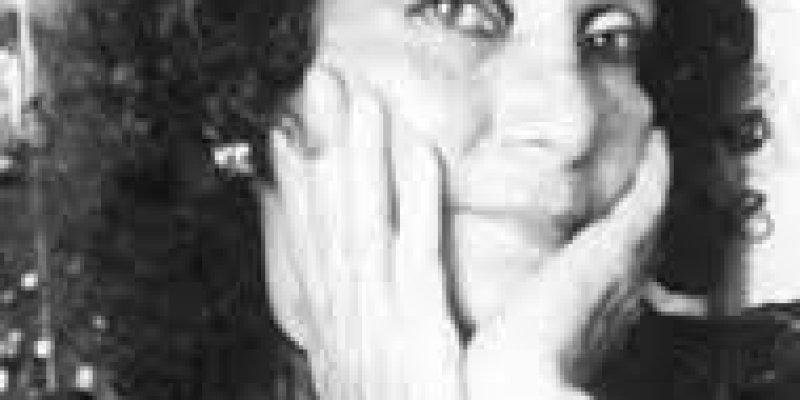 Sevim Burak’ın siyah-beyaz yakın plan bir portre fotoğrafı. Kıvırcık, koyu renk saçlı, her iki eli yüzüne dayalı; avuç içleri yanaklarda, parmaklar şakaklara doğru uzanıyor. Üzerinde parlak görünümlü koyu renk bir üst var. kameraya doğru bakıyor ve gülümsüyor. Arka plan kalabalık.