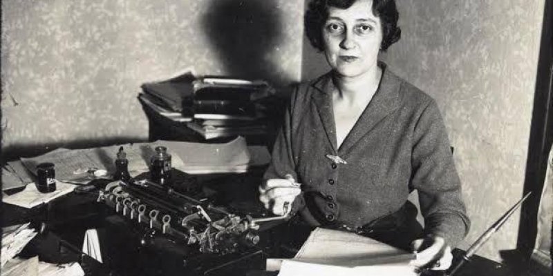 Selma Rıza bir masanın arkasında oturuyor. Kısa ve dalgalı saçlı ve ciddi bir ifadeyle kameraya bakıyor. Üzerinde düğmeli, koyu renkli bir hırka var. Masanın üzerinde bir daktilo, birçok kâğıt ve belgeler bulunuyor. Selma Rıza elinde bir kalem tutuyor ve önündeki kâğıtlarla meşgul. Masanın arka tarafında üst üste yığılmış daha fazla kâğıt ve dosya var. Oda, sade bir duvar kâğıdıyla kaplı.