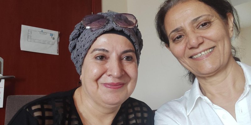 Solda gri bonesi üzerinde pembe güneş gözlüğü ve siyah bluzu ile bir kadın var. Sağda beyaz, sade gömleği, toplu saçları olan kadın gülüyor.