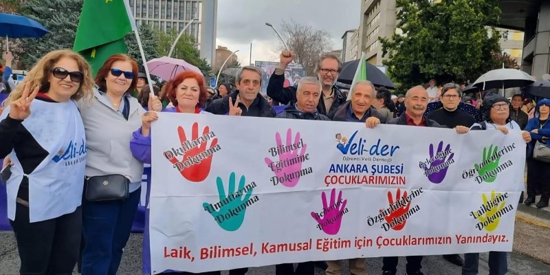 Kalabalık bir protesto yürüyüşünde pankart tutan bir grup yetişkin. Önde farklı yaşlarda kadın ve erkeklerden oluşan yaklaşık on kişilik bir grup var; çoğu gülümsüyor, bazıları zafer işareti yapıyor, biri yumruğunu havaya kaldırmış. En soldaki kadın sarı dalgalı saçlı, güneş gözlüklü, beyaz önlük giymiş; yanında kısa kırmızı saçlı başka bir kadın, onun yanında kırmızı saçlı, elinde pankartın sol kenarını tutan bir kadın duruyor. Sağ tarafta gri saçlı ve sakallı erkekler, aralarında gözlüklü bir
