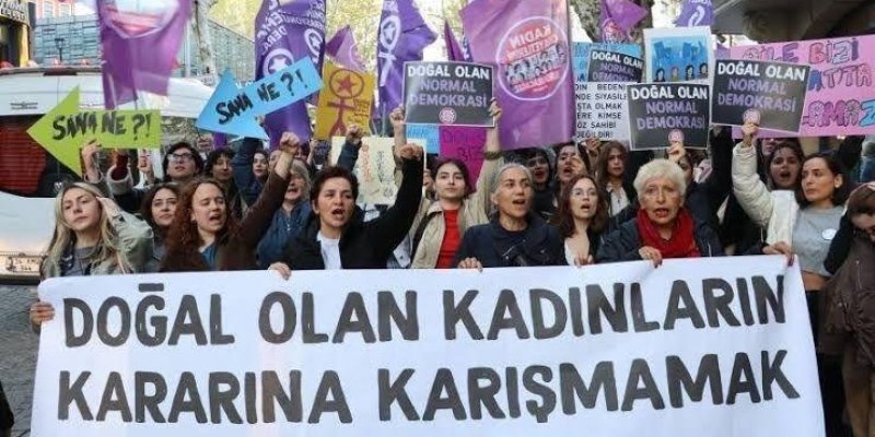 Kalabalık bir grup kadın, bir protesto yürüyüşünde ellerinde pankartlar ve dövizler taşıyor. En önde büyük bir pankartta "DOĞAL OLAN KADINLARIN KARARINA KARIŞMAMAK", arkadaki dövizlerde "DOĞAL OLAN NORMAL DEMOKRASİ", "ŞİMDİ NE?!" gibi ifadeler yazıyor. Kadınlar çeşitli yaş gruplarından ve hepsi kararlı bir şekilde slogan atıyor. Arka planda mor ve pembe bayraklar dikkat çekiyor.