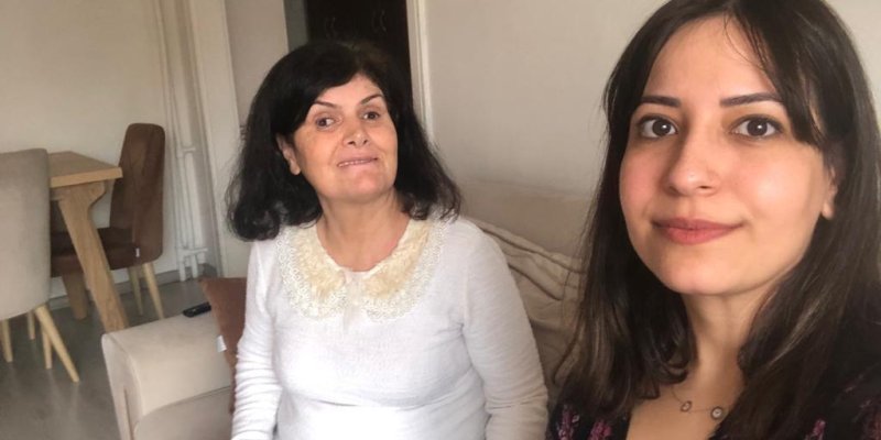 Kanepede yan yana oturan iki kadından kameraya yakın olan genç kadın uzun, koyu kahve saçları ve buğday teniyle hafifçe tebessüm ediyor, arkada Şule Sepin İçli koyu renk omuz hizasındaki saçları, beyaz kazağıyla gülümsüyor.