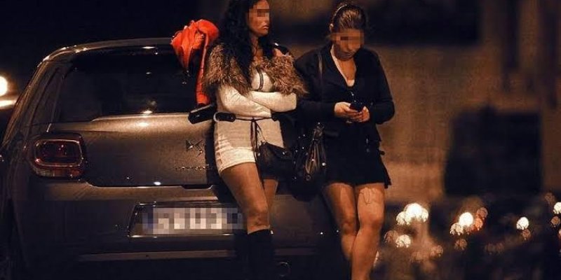 Gece vakti bir sokakta park etmiş bir arabanın yanında duran iki kadın. Biri beyaz elbise ve siyah çizmeler giymiş, diğeri siyah elbise giymiş. Her iki kadının da yüzleri bulanıklaştırılmış. Arabanın arka planında sokak lambalarının ışıkları ve bulanık bir şehir manzarası var. Kadınlardan biri telefonuna bakıyor, diğeri kollarını göğsünde kavuşturmuş şekilde duruyor. Arabanın üzerinde kırmızı bir ceket asılı.