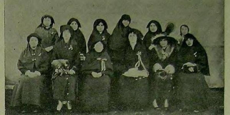 Eski bir siyah-beyaz grup fotoğrafı. İki sıra halinde oturan ve ayakta duran on bir kadın var. Geleneksel kıyafetler giymişler ve çoğu başörtüsü takıyor. Ön sırada oturan kadınlardan biri büyük bir şapka takıyor. Fotoğrafın alt kısmında, Farsça bir yazı bulunuyor.
