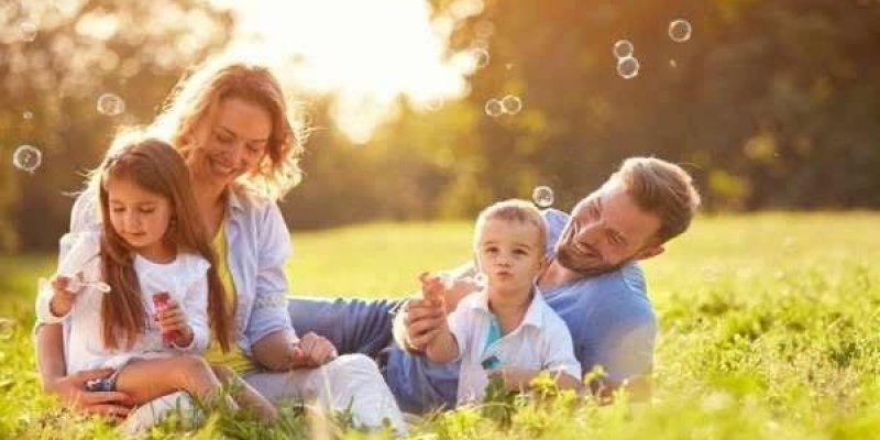 Güneşli bir günde çimenlerin üzerinde oturan mutlu bir aile. Anne, baba ve iki çocuk var. Anne ve baba, çocuklarıyla birlikte sabun köpükleri üflüyorlar. Anne, uzun sarı saçlı ve beyaz bir gömlek giymiş. Baba, kısa saçlı ve mavi bir gömlek giymiş. Küçük kız, beyaz bir elbise giymiş ve uzun kahverengi saçları var. Küçük oğlan çocuk beyaz bir tişört giymiş. Herkes gülümsüyor. Arka planda ağaçlar ve güneş ışığı.