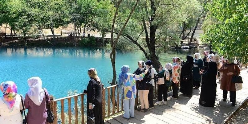 Bir grup kadın ahşap bir platform üzerinde durarak mavi renkli bir gölü izliyor. Kadınların çoğu başörtüsü takıyor ve çeşitli renklerde kıyafetler giymişler. Platformun etrafında ahşap bir korkuluk var ve arka planda ağaçlar görülüyor. Gölün suyu berrak ve etrafı yeşilliklerle çevrili. Hava açık ve güneşli.