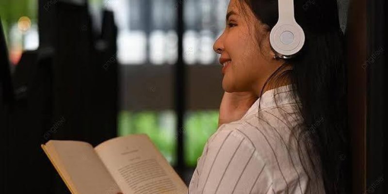 Beyaz kulaklık takmış bir kadın, bir duvara yaslanmış ve elinde açık bir kitap tutuyor. Gülümsüyor ve rahat görünüyor. Arka planda bulanık bir pencere ve dışarıdaki yeşillikler seçiliyor. Kadın beyaz çizgili bir gömlek giymiş.