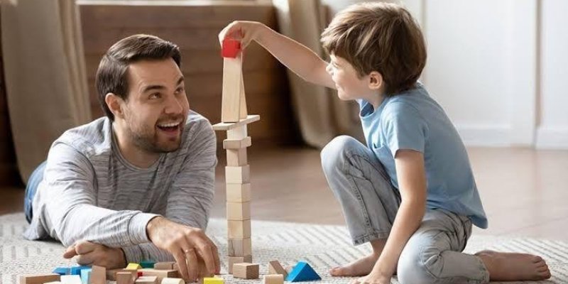 Bir adam ve küçük bir çocuk yerde oturmuş, birlikte ahşap bloklarla oynuyorlar. Adam yerde uzanmış, gülümseyerek çocuğa bakıyor. Çocuk dizlerinin üzerinde oturmuş, elinde kırmızı bir blok tutuyor ve blokları üst üste dizerek yüksek bir kule yapmaya çalışıyor. İkisi de mutlu ve eğlenceli bir an yaşıyor. Arka planda, odanın bir köşesi ve pencere perdeleri görünüyor.