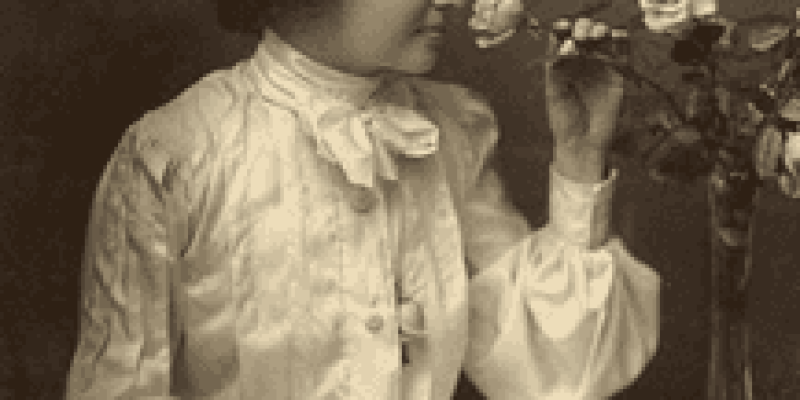 Helen Keller’in 20 li yaşlarına ait siyah beyaz fotoğrafında Keller sandalyeye oturmuş, beyaz uzun kollu bir gömlek giymiş, saçlarını alttan topuz yapmış. Dizlerinin üzerinde büyük sayfaları açık bir defter var. Elinin teki defterin üzerinde duruyor. Diğer eliyle de yan tarafında sehpada duran vazodaki güllere uzanmış ve güller kokluyor.