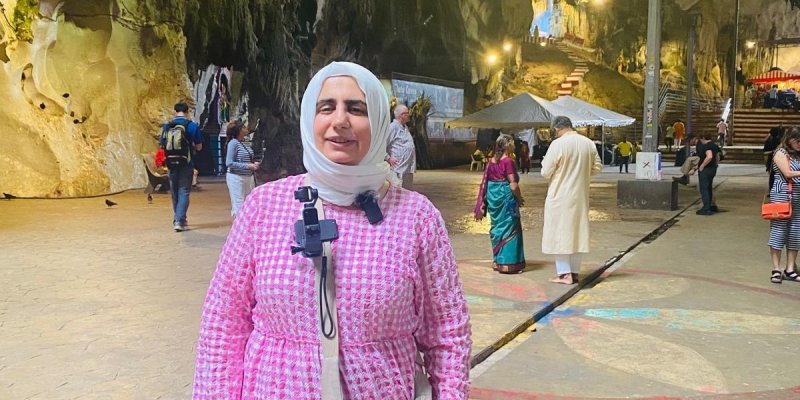 Pembe ve beyaz desenli uzun bir elbise giyen, beyaz başörtüsü takan Esma Gökalp. Boynunda bir kamera asılı. Omzunda açık gri renkte bir çanta taşıyor. Arka planda büyük bir mağara içindeler ve mağaranın duvarları doğal taş yapısıyla dikkat çekiyor. Mağaranın içinde başka insanlar da var ve bazıları yürürken bazıları duruyor. Mağaranın içi aydınlatılmış ve ileride merdivenler görülüyor. Çeşitli renklerde giysiler giymiş insanlar mağaranın içinde dolaşıyor.
