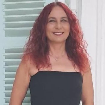 Kıvırcık kızıl saçlı, orta tenli. Açık renkli panjurlu bir pencerenin önünde ayakta duruyor. Saçları omuzlarının biraz altına kadar iniyor ve doğal dalgalı. Yüzünde hafif bir makyaj; dudakları doğal bir tonda, kaşları belirgin ve yüz ifadesi sakin, gülümsüyor. Askısız, düz kesim, siyah bir elbise giymiş; elbise göğüs üstünden düz bir hatla başlıyor ve üst kısmı vücuda oturmuş. Boynunda ince bir zincir kolye ve küçük bir kolye ucu.
