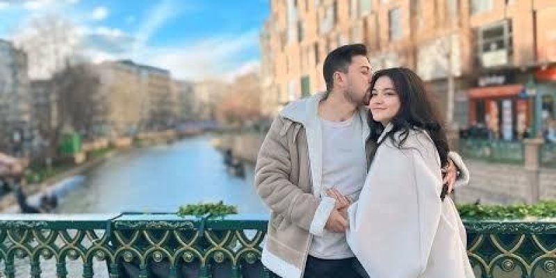 Köprü korkuluğunun önünde birbirine sarılmış duran bir çift. Erkek solda, kadın sağda duruyor; erkek kadının başının yanından öpüyor. Erkek kısa koyu saçlı, teni açık; bej renkli kalın bir ceket ve açık renk bir üst, koyu pantolon ve beyaz spor ayakkabı giymiş. Kadın uzun koyu saçlı, teni açık; beyaz geniş bir üst ve koyu pantolon, koyu renk ayakkabı giymiş. Arka planda kanallı bir su yolu, suyun iki yanında binalar ve ağaçlar, mavi gökyüzü ve bulutlar. Köprü korkuluğu yeşil renkte, üst kısmında