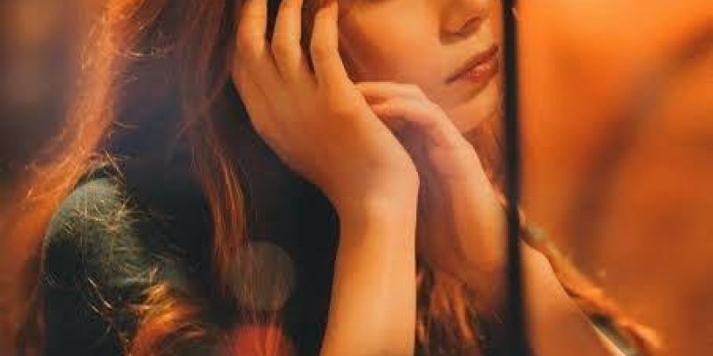 Turuncu-sıcak ışıkta bir kafede masada oturan genç bir kadın portresi. Uzun, dalgalı kızıl saçlı ve açık tenli; başını hafif yana çevirmiş, uzak bir noktaya dalgın bir ifadeyle bakıyor. Dirsekleri masada, iki eliyle yüzünü/çenesini destekliyor. Üzerinde koyu renkli uzun kollu bir üst var. Masanın ön kısmında bulanık bir saksı bitkisi ve yanında küçük bir cam kap. Arka planda yuvarlak ışıklar ve kahverengi-turuncu tonlarda bulanık bir ortam.