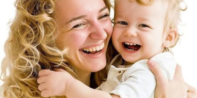 Kıvırcık sarı saçlı bir yetişkin kadın ile kıvırcık açık sarı saçlı küçük bir çocuk, yakın planda sarılarak gülüyor. Kadının teni açık ve yüzü yana dönük; gözleri kısık şekilde gülümsüyor. Çocuğun teni açık, ağzı açık kahkaha atıyor ve kadına kollarını dolamış. İkisinin de üstünde beyaz tonlarda kıyafetler var. Arka plan düz beyaz.