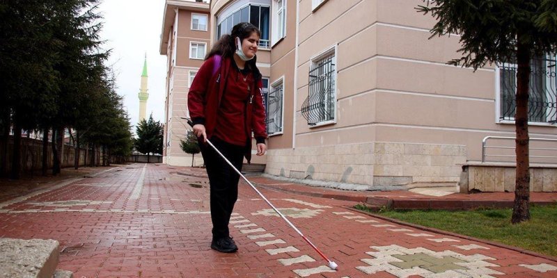 Kırmızı tuğla döşeli bir yürüyüş yolunda beyaz bastonla yürüyen bir kişi. Üzerinde bordo bir mont, siyah pantolon ve sırt çantası var; bastonunu önünde yere doğru uzatmış. Sağ tarafta bej renkli, pencerelerinde demir parmaklıklar olan bir bina uzanıyor; bina kenarında küçük bir yeşil alan ve ağaç görülüyor. Sol tarafta ağaç sırası boyunca uzayan bir kaldırım ve arka planda yeşil sivri kubbeli bir minare seçiliyor.