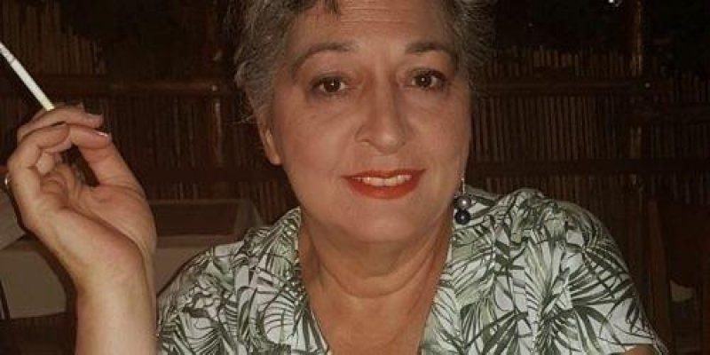 Orta yaşlarda kısa saçlı bir kadın beyaz üzerine yeşil yapraklı bluzu, sağ elinde tuttuğu sigarasıyla gülümsüyor.