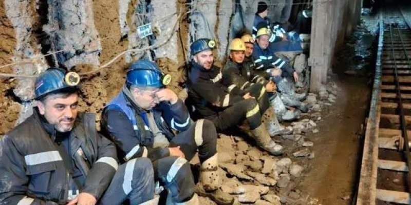 Maden ocağında dinlenen işçiler. Sarımsı-kahverengi, nemli ve parçalanmış kaya yüzeyinin yanında yere oturmuş altı madenci. Hepsinin üzerinde koyu renkli, yansıtıcı şeritli iş kıyafetleri ve açık renk, dizlere kadar uzanan lastik çizmeler; kıyafetleri ve çizmeleri çamur ve toz içinde. Başlarında lambalı baretler; ön taraftaki dört madencinin baretleri mavi, ortalarda bir işçinin bareti sarı, en arkadakilerden birinde koyu renk baret. Madenciler tünelin sol duvarına sırtlarını yaslamış, aralarınd