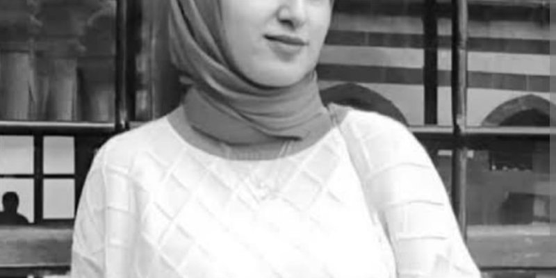 Pencereli/metal parmaklıklı bir arka plan önünde duran Rojin Kabaiş’in portresi. Fotoğraf siyah-beyaz. Başını hafifçe yana çevirmiş ve kadrajın sağına doğru bakıyor. Yüz ifadesi sakin, hafif bir gülümseme var. Ten rengi açık. Başında düz, koyu tonlu bir başörtüsü, boyun kısmını da sarıyor. Üzerinde açık renk, baklava desenli bir kazak, omuzdan çapraz geçen ince bir çanta askısı. Arka planda büyük pencere bölmeleri ve metal ızgara çizgileri belirgin.