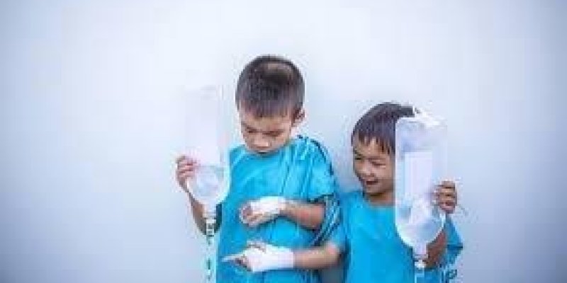 5-6 yaşlarında biri kız iki çocuk mavi renkli hasta kıyafeti giyinmişler. Kollarında serum takılı halde ayakta duruyorlar.