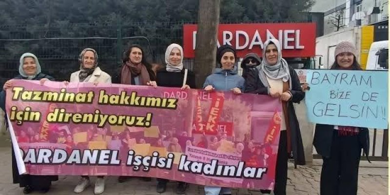 Kaldırım kenarında yan yana duran bir grup kadın ve ellerinde protesto pankartları. Kadınlar dış mekânda, bir fabrikanın ya da tesisin girişine benzer bir noktada duruyor; arkada tel çit ve büyük kırmızı bir tabela üzerinde “DARDANEL” yazısı. Grup yaklaşık 9 kişiden oluşuyor. Çoğu başörtülü; bir kişi güneş gözlüğü takmış. Kıyafetler kışlık görünümlü (mont, uzun kaban, hırka gibi). Ön tarafta pembe tonlarda büyük bir pankart tutuluyor; pankartın üzerinde kalabalık fotoğrafı ve kırmızı bayrak görs