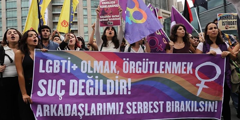 Renkli bir pankart taşıyan kalabalık bir protesto grubu. Pankart mor zeminli, ortasından gökkuşağı renklerinde dalgalı bir şerit geçiyor ve üzerinde büyük beyaz harflerle: “LGBTI+ OLMAK, ÖRGÜTLENMEK SUÇ DEĞİLDİR! ARKADAŞLARIMIZ SERBEST BIRAKILSIN!” yazısı. Sağ tarafta kadınlık sembolü beyaz renkte, içi gökkuşağı renkleriyle dolu. Pankartın arkasında çoğu genç yetişkinlerden oluşan bir grup; ağızları açık, slogan atıyorlar.