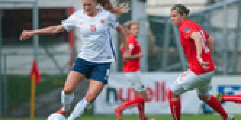 Futbol sahasında futbol oynayan 3 kadın var. Geride duran iki kadın kırmızı uzun çorap, kırmızı kısa kollu forma, beyaz şort giyinmiş. Topu ayağında tutan ve önde duran üçüncü kadın beyaz kısa kollu tişört ve uzun çorap, lacivert şort ve kırmızı spor ayakkabı giymiş. Kadın uzun düz sarı saçlı, saçları atkuyruğu şeklinde arkadan bağlı.