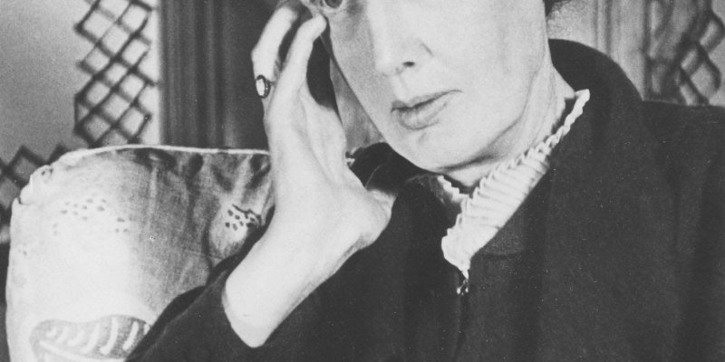 Ünlü İngiliz yazar Virginia Woolf. Siyah-beyaz bir portre fotoğrafı. Woolf, başını eline yaslamış bir şekilde düşünceli bir ifadeyle kameraya bakıyor. Saçları geriye doğru taranmış ve sade bir kıyafet giymiş. Arka planda, desenli bir yastık ve ahşap bir mobilya parçası görülüyor.
