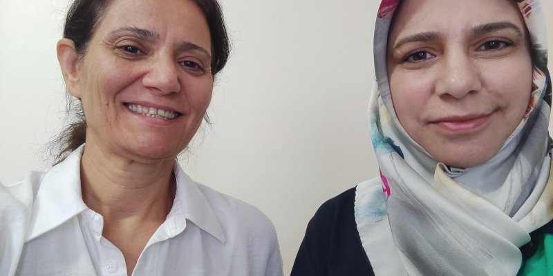 İki kadın omuz omuza duruyor. Sol taraftaki kadın beyaz bir gömlek giymiş, saçları kahverengi ve omuzlarına kadar uzunlukta. Gülümsüyor.