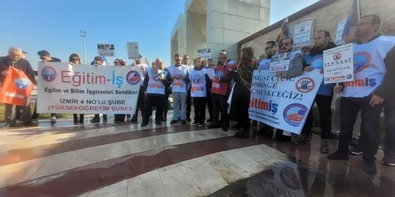 Bir grup insan ellerinde pankartlar tutuyor. Pankartların üzerinde "Eğitim-İş Eğitim ve Bilim İşgörenleri Sendikası" yazıyor. "İzmir 4 No'lu Şube (Yükseköğretim Şubesi)" ifadesi de yer alıyor. Diğer pankartlarda "Angusluk, Mobbing Geçmeyeceğiz!" ve "12.5 Saat Angarya Olur" gibi yazılar bulunuyor. Katılımcılar beyaz tişörtler giymiş ve bazıları güneş gözlüğü takıyor.