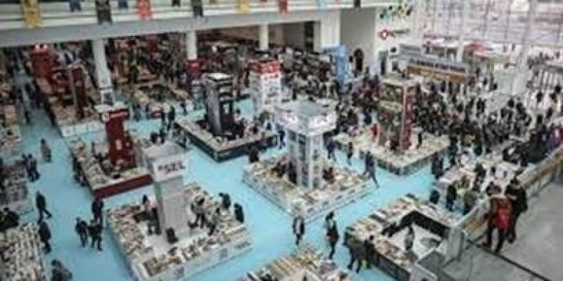 Açık mavi zeminli büyük kapalı bir ortamda kare kare oluşturulmuş kitap stantlarının tepeden çekilmiş görüntüsü var. Stantların üzerinde kitaplar dizilmiş. Stantların etrafında birçok insan görünüyor.