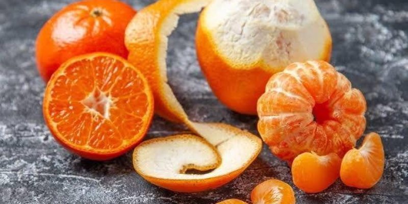 Bir grup mandalina. Bir tanesi tamamen soyulmuş ve dilimlere ayrılmış. Bir diğeri kısmen soyulmuş, kabuğu hala üzerinde duruyor.Bir mandalina da ortadan ikiye kesilmiş ve iç kısmı görünüyor. Arka planda soyulmamış bir mandalina var. Mandalinaların kabukları parlak turuncu renkte ve yüzeyleri pürüzsüz. Gri ve dokulu bir yüzeyi olan zemin.