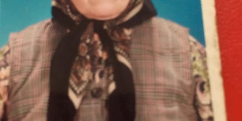 65-70 yaşlarında bir kadının stüdyoda çekilmiş bir fotoğrafı. Kadın beyaz tenli, başında kahverengi bir baş örtüsü var. Toprak rengi bir yelek giymiş, yeleğin içinde kahverengi tonlarında çiçekli uzun kollu bir bluz var.