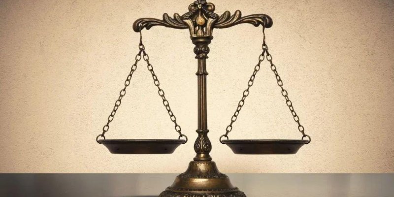 Eski tarz bir terazi heykeli. Ortada ince, yukarı doğru uzanan ve üzerinde oymalı süsler bulunan bronz renkli bir sütunun tepesinden iki yana doğru kıvrılan dekoratif kollar çıkıyor. Bu kollardan aşağıya doğru ince zincirler sarkıyor ve zincirlerin ucunda yuvarlak, sığ iki terazi tabağı asılı. Sağ ve sol tabak aynı hizada, dengede duruyor. Alt kısımda geniş, oymalı desenli, yuvarlak bir kaide. Arka planda açık bej tonlarında düz bir duvar, altında nötr renkli düz bir zemin.