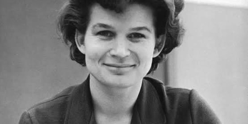Orta yaşlarında, beyaz tenli Valentina Tereşkova’nın portresi. Omuz hizasından yukarıya kadar görülüyor ve doğrudan kameraya doğru hafifçe gülümseyerek bakıyor. Kısa, dalgalı, kabarık bir saç modeli var; saçları koyu renk ve alnına doğru hafifçe dökülmüş. Yüz hatları yumuşak, dudakları kapalı biçimde gülümsüyor, gözleri koyu renk. Üzerinde yakalı, tek renk ceket koyu tonda. Ceketin sol tarafında küçük dikdörtgen bir rozet ve altında yıldız şeklinde büyükçe bir madalya asılı. Arka plan sade ve bu