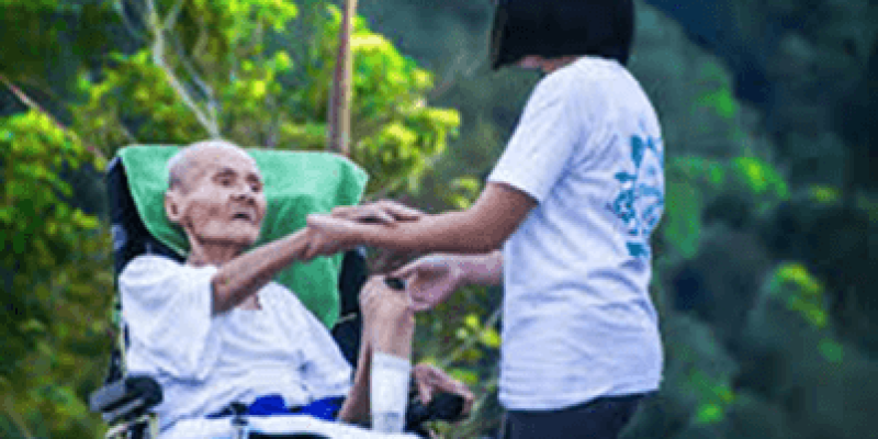 Tekerlekli sandalyede oturan yaşlı bir adam ve ona yardım eden genç bir kadın var. Adam beyaz bir gömlek, kadın siyah bir tişört giymiş. İkisi de birbirlerine bakıyor ve kadın, adamın ellerini tutuyor. Arkalarında ağaçlar ve yeşillikler bulunuyor.