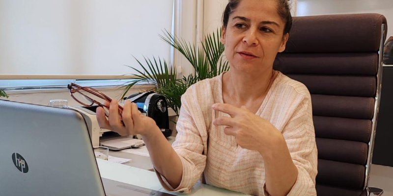 Açık tenli, orta yaşlardaki Özlem Ayata, ofis ortamında çalışıyor. Açık bej rengi bir bluz giymiş ve saçları toplanmış. Masasında bir dizüstü bilgisayar, birkaç defter ve kalem var. Bir elinde telefon tutarken diğer eliyle bir şeye işaret ediyor gibi. Arka planda bir pencere, birkaç saksı bitki ve bir tablo bulunuyor. Tabloda, deniz kenarında oturan iki kişi tasvir edilmiş.