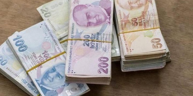 Masanın üzerinde üç farklı banknot destesi. Solda 100 Türk Lirası banknotlarından oluşan bir deste var. Ortada 200 Türk Lirası banknotlarından oluşan bir başka deste bulunuyor. Sağda 50 Türk Lirası banknotlarından oluşan bir deste yer alıyor. Her bir deste, lastik bantlarla sıkıca bağlanmış. Banknotların üzerinde Mustafa Kemal Atatürk'ün portresi bulunuyor. Arka plan ahşap bir yüzey.