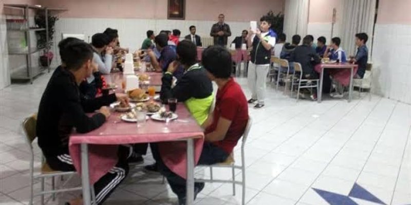 Bir yemekhanede yemek yiyen bir grup genç erkek. Uzun bir masa etrafında oturmuşlar ve yemeklerini yiyorlar. Masada tabaklar, bardaklar ve içecekler var. Arka planda, ayakta duran bir kişi bir şeyler anlatıyor. Yemekhanenin zemininde beyaz fayanslar ve mavi desenler var. Duvarlar açık renkli ve bir köşede bitki yer alıyor.