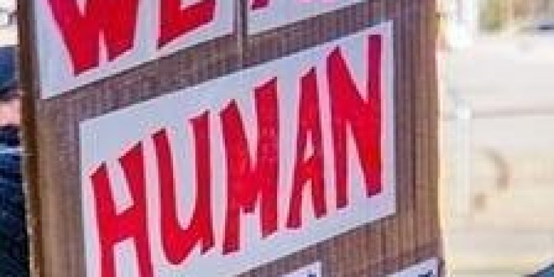 Kahverengi zeminde kırmızı büyük harflerle “We are human” yazılı pankart var.