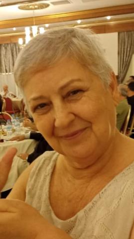 Yakın çekim fotoğraf. Kısa, gri saçları var ve hafif gülümseyerek kameraya bakıyor. Üzerinde krem veya açık renkli, dantel desenli bir bluz bulunuyor. Arka planda bir restoran ya da davet salonu gibi bir ortam görünüyor; masada su şişeleri duruyor ve diğer insanlarla dolu yuvarlak masalar bulunuyor. Ortamda sıcak bir ışık hakim.